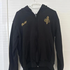 Bosnien hoodie - Svart hoodie med huva och dragkedja, dekorerad med guldfärgade broderier fram och bak. På bröstet finns en fleur-de-lis och texten 'BiH', medan ryggen har ett stort broderat emblem med texten 'Jedna si Jedina'. Mjuk bomullskänsla och praktiska fickor framtill.