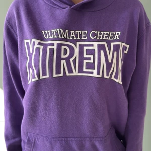 Lila hoodie Ultimate Cheer Xtreme - Säljer en lila hoodie från Ultimate Cheer Xtreme med stor vit text på bröstet. Tröjan har huva med snörning, magficka och långa ärmar. Perfekt för dig som gillar cheer eller vill ha en chill och färgglad look.