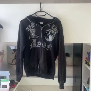 Svart hoodie med strasstenar och tryck - Svart hoodie med dragkedja från Aim for the moon. Framsidan har ett stort tryck med djävulsmotiv och text i strasstenar. Huva och fickor framtill. Perfekt för dig som gillar streetstyle och vill sticka ut med bling. Säljer för jag vill köpa i en annan färg 💓 pris kan diskuteras 