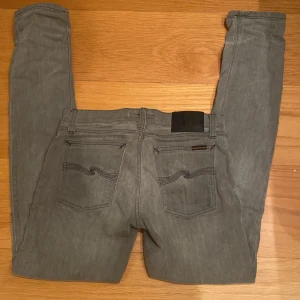 Grå skinny jeans från Nudie Jeans - Snygga grå jeans från Nudie Jeans med klassisk femficksdesign och svarta detaljer på bakfickorna. Modellen är skinny med smal passform och normal midja. Jeansen är tillverkade i mjukt jeanstyg och har svarta sömmar och Nudie-logga på bakfickan.