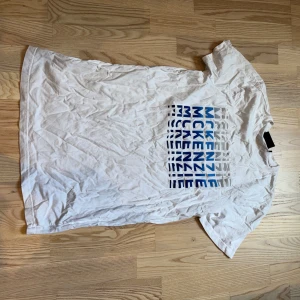 Vit McKenzie t-shirt med blått tryck - Vit t-shirt från McKenzie med coolt grafiskt tryck i blått och grått på bröstet. Klassisk rund hals och korta ärmar. Materialet är mjuk bomull, perfekt för chill dagar. Snyggt streetwear-plagg med relaxed vibe.