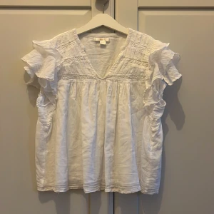 Vit blus med volangärmar Gina Tricot - Supersöt vit blus från Gina Tricot med v-ringning och volangärmar. Blusen har broderade detaljer upptill och är tillverkad i ett luftigt bomullstyg. Perfekt för dig som gillar romantisk och chill stil. Volangerna ger extra flärd och gör toppen unik.