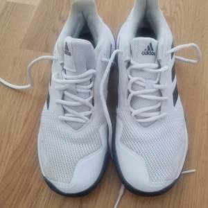 Vita Adidas sneakers med svarta ränder - Fräscha vita Adidas sneakers med klassiska svarta ränder på sidan. Skorna har mesh-ovandel för bra ventilation, snörning och en platt sula med tydligt mönster för extra grepp. Perfekta för dig som gillar sportig stil och vill ha bekväma skor till träning eller häng.