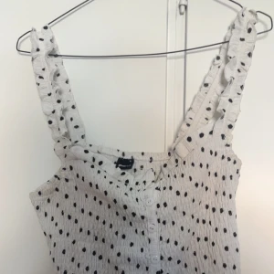 Prickig vit linnetopp med volang - Supersöt vit linnetopp från Gina Tricot med svarta prickar, knappar framtill och breda axelband med volangdetaljer. Toppen har en smockad, stretchig passform och volangkant nedtill. Den är rätt så liten i storleken, jag brukar bära S.