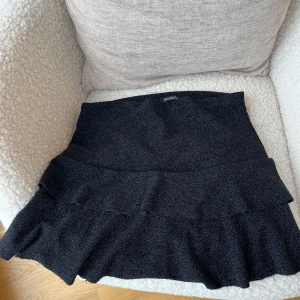 Svart volangkjol från Missay - Supersnygg svart kortkjol från Missay med volangdetaljer. Kjolen har en tight passform upptill och är tillverkad i ett glittrigt syntetmaterial som ger en cool textur. Perfekt för dig som vill sticka ut med en trendig look. OBS på grund av materialet så sticker det ut trådar från den. Se bild 2 för att se ! 