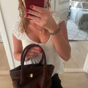 Brun mocka handväska med gulddetalj - Snygg brun handväska i mjuk mocka med stilren guldfärgad låsdetalj framtill. Väskan har två rejäla handtag och en klassisk, lite boxig form som gör den enkel att bära med sig. Perfekt storlek för det viktigaste och ger en cool vibe till din outfit.