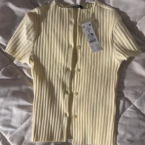 Ribbad ljusgul topp från Gina Tricot - Säljer en ribbad ljusgul topp från Gina Tricot med korta ärmar och knappar hela vägen framtill. Sitter tyvärr för tajt på mig därav säljer jag den. Toppen har en snygg, vertikal ribbad struktur och är tillverkad i mjuk bomullsmix. 