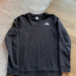 Svart sweatshirt från The North Face - Säljer en svart sweatshirt från The North Face med diskret logga på bröstet. Tröjan har rund hals, långa ärmar och ribbade muddar vid ärmslut och nederkant. Perfekt för chill dagar eller när du vill ha en enkel och stilren look.