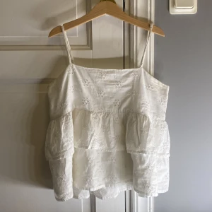 Vit blommig linnetopp från Gina Tricot - Supersöt vit linnetopp från Gina Tricot med tunna axelband och broderade små blommor. Toppen har volanger i två lager som ger en luftig och chill vibe. Perfekt för varma dagar och enkel att matcha med jeans eller shorts.