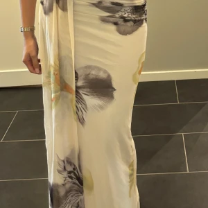 Lång blommig klänning i ljusa toner - Säljer en elegant långklänning med blommigt mönster i ljusa färger som vitt, grått och inslag av beige och gul. Klänningen har omlott-detalj och är tillverkad i ett mjukt, lätt material som känns svalt mot huden. Perfekt för sommarens alla tillfällen. Köpt i Grekland