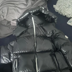 Svart Moncler pufferjacka - Säljer en svart pufferjacka från Moncler med glansig finish och stor huva. Jackan har tydliga quiltade partier, dragkedjor i silver och Moncler-logga på ärmen. Perfekt för kalla dagar och har en riktigt schysst streetstil.
