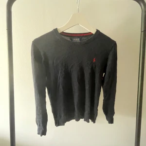 Mörkblå tröja från Polo Ralph Lauren - Mörkblå långärmad tröja från Polo Ralph Lauren i mjuk pima bomull. Klassisk rund hals och röd broderad logga på bröstet. Perfekt för dig som gillar stilrena och tidlösa plagg med en touch av lyx. Passar dig som vill ha en clean look med diskreta detaljer.140/152