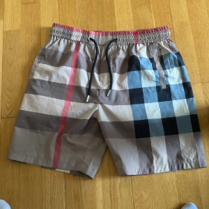 Burberry shorts - Snygga Burberry shorts med klassiskt rutigt mönster i beige, svart, blått och rött. De har elastisk midja med svart snörning och fickor både fram och bak. Perfekta för sommaren och har en loose passform som ger chill vibe. Aldrig använda.