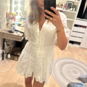 Vit kort klänning med spets och bälte - Superfin vit kort klänning med broderad spets och knytbälte i midjan. Klänningen har v-ringad hals, knappar framtill och är kortärmad. Perfekt för sommarens alla tillfällen, med luftig bomullskänsla och snygg volang nedtill.