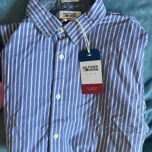 Tommy Hilfiger Skjorta - Snygg blå skjorta med vita vertikala ränder från Tommy Hilfiger Denim. Klassisk krage, långärmad och med vita knappar framtill. Ikonisk Hilfiger-logga broderad på bröstet. Perfekt för dig som gillar stilrena och fräscha plagg. Gåva av min bror men den var för stor.