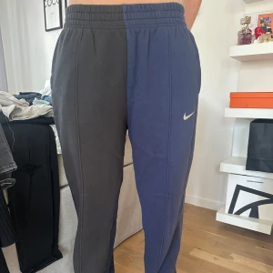 Nike tvåfärgade mjukisbyxor S - Säljer ett par unika mjukisbyxor från Nike i storlek S. Ena benet är mörkgrått och det andra är mörkblått, med klassisk Nike-logga på höger ben. Byxorna har elastisk midja och raka ben, perfekta för chill eller träning. Grym stil för dig som gillar att sticka ut.