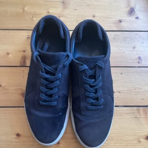 Axel Arigato skor - Snygga svarta sneakers i mocka med klassisk låg profil och vit platt sula. Skorna har rund tå och snörning framtill för en clean look. Perfekta för dig som gillar stilrena och enkla sneakers med en mjuk känsla.