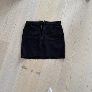 Svart jeanskjol från Carin Wester - Svart kort jeanskjol från Carin Wester med rå kant nertill och klassiska fickor fram och bak. Kjolen har hällor för bälte och stängs med knapp och dragkedja. Perfekt för en cool och avslappnad stil.