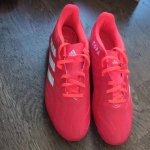 Adidas Copa röda fotbollsskor - Snygga Adidas Copa fotbollsskor i en intensiv röd färg med vita detaljer och klassiska Adidas-ränder på sidan. Skorna har snörning och en platt sula med röda och vita dobbar för grymt grepp på planen. Ovandelen är i syntetmaterial och designen är sportig och modern.