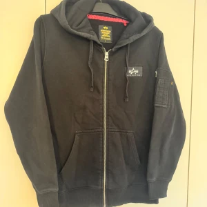 Svart hoodie Alpha Industries M - Svart hoodie från Alpha Industries med stor vit logga på ryggen och mindre logga på bröstet. Tröjan har huva med snörning, dragkedja framtill, fickor och en extra ficka med dragkedja på ärmen. Mjuk insida och ribbade muddar. Perfekt för chill eller streetwear.
