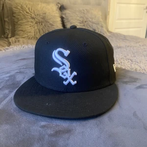 Svart New Era 59FIFTY White Sox keps, profil för mer - Säljer en svart New Era 59FIFTY keps med Chicago White Sox-logga i vitt framtill och MLB-logga baktill. Klassisk flat peak och broderade detaljer. Perfekt för dig som gillar sportig streetstyle och vill ha en snygg accessoar med lagkänsla.