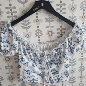 Vit blommig croppad blus med snörning - Superfin vit blus med blått blommigt mönster och puffiga korta ärmar. Toppen är croppad och har en söt snörning framtill samt volangkant vid halsen. Perfekt för dig som gillar romantisk och trendig stil.