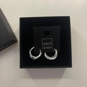 Silverörhängen från SNÖ of Sweden - Snygga örhängen i silverfärg från SNÖ of Sweden. De är formade som tvinnade hoops med glittrande detaljer på ena sidan som ger extra bling. Perfekta för att lyfta din stil med en modern och elegant touch.