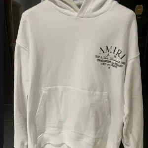 Vit hoodie från Amiri med tryck - Snygg vit hoodie från Amiri med svart texttryck både fram och bak. Tröjan har huva, känguruficka och avslappnad passform. Perfekt för dig som gillar streetwear och vill sticka ut med ett stilrent plagg.