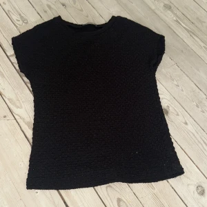 Svart stickad topp från Zara - Svart stickad topp från Zara med korta ärmar och rund hals. Materialet har en grov, texturerad yta som ger en cool och unik känsla. Perfekt att styla med jeans eller kjol för en chill look. Enkel och stilren design som funkar året runt.