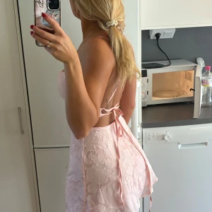 Ljusrosa kort klänning med snörning - Säljer en superfin ljusrosa kort klänning med tunna axelband och öppen rygg med snörning. Klänningen har ett blommigt mönster i tyget och är figurnära med omlottkänsla baktill. Perfekt för sommarens alla tillfällen!