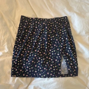 Blommig mörkblå kortkjol med slits - Supersöt mörkblå kortkjol med små rosa och gröna blommor över hela tyget. Kjolen har en liten slits framtill som ger en extra touch. Perfekt för dig som gillar romantiska och lekfulla detaljer i din stil.