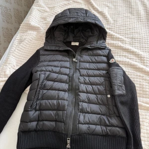 Svart Moncler pufferjacka med huva - Snygg svart pufferjacka från Moncler med stickade ärmar och huva. Jackan har quiltad front, dragkedja och Moncler-logga på ärmen. Perfekt för dig som gillar streetstyle och vill ha något varmt och trendigt. Flera fickor och ribbade muddar för extra komfort.