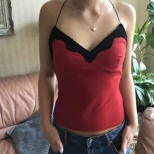 Röd linnetopp med svarta spetsdetaljer - Snygg röd linnetopp med tunna axelband och svarta spetsdetaljer längs bysten. Toppen har en feminin v-ringning och är figurnära med en mjuk, slät yta. Perfekt för dig som vill ha en trendig och stilren look med lite extra edge.