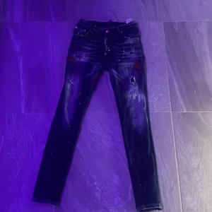 Dsquared2 blå jeans med röda detaljer - Snygga blå jeans från Dsquared2 med coola slitningar, färgstänk och röda broderade lönnlöv på benen. Jeansen har en rak passform, klassisk femficksdesign och tydliga vita och röda detaljer som ger en unik streetkänsla. Perfekt för dig som gillar statement-plagg.