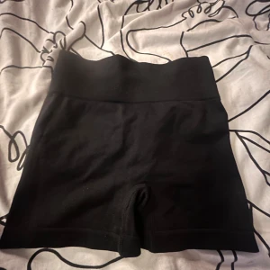 Svarta högmidjade träningsshorts XS - Säljer ett par svarta högmidjade träningsshorts i storlek XS. De är tillverkade av stretchigt material med 92% polyamid och 8% elastan, vilket gör dem riktigt bekväma och följsamma. Baktill har de snygg rynkdetalj som framhäver formen.