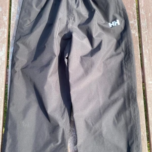 Svarta täckbyxor från Helly Hansen - Svarta täckbyxor från Helly Hansen med Helly Tech Protection-tyg som är både vind- och vattentätt. Byxorna har en loose passform och justerbara kardborreband vid bensluten. Perfekta för kalla och blöta dagar utomhus.