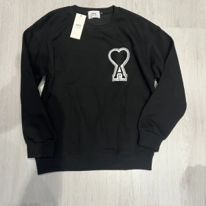 Svart sweatshirt från Ami Paris - Svart sweatshirt från Ami Paris med stort broderat hjärta och bokstaven A på bröstet. Tröjan har rund hals, ribbade muddar och är tillverkad i mjuk bomull. Perfekt för dig som gillar stilrena och coola detaljer.