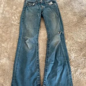 Blå bootcut jeans från Gina Tricot - Snygga blå jeans från Gina Tricot i bootcut-modell med klassisk femficksdesign och dubbla knappar i midjan. Jeansen har en ljus tvätt och är tillverkade i jeansmaterial med kontrastsömmar. De har några få fläckar som knappt syns. Midjemått - 32 cm innerbenslängd- 76 cm