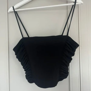 Svart korsett-topp från Zara - Svart korsett-topp från Zara med smala axelband och snygga rynkade sidor som ger en cool och trendig look. Toppen har en croppad passform och är tillverkad i ett mjukt polyestermaterial. Perfekt för dig som vill sticka ut med en edgy vibe.