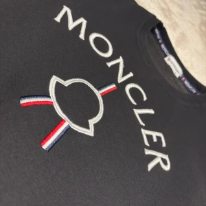 Svart Moncler sweatshirt - Säljer en tvär snygg svart sweatshirt från Moncler med stort broderat logotyp på bröstet i vitt, rött och blått. Tröjan har rund hals, långa ärmar och ribbade muddar vid ärmslut och nederkant. Vid frågor så är de bara att höra av sig!