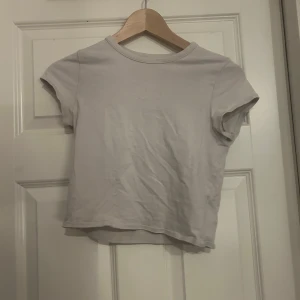 Ljusvit croppad t-shirt - En enkel och fräsch ljusvit croppad t-shirt med rund hals och korta ärmar. Toppen har en clean look och är gjord i mjuk bomull, perfekt för att matcha med jeans eller kjol. Snygg passform som sitter lite tight och ger en modern vibe.