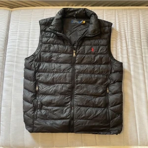 Ralph lauren väst - Svart dunväst från Polo Ralph Lauren med quiltad design och ståkrage. Västen har dragkedja framtill, två sidofickor med dragkedja och en röd broderad logga på bröstet. Perfekt att slänga över en hoodie när det är kyligt ute.