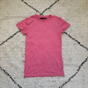 Rosa ribbad t-shirt från lager 157 - Snygg rosa t-shirt från lager 157 i storlek XS. Modellen är ribbad och har en klassisk rund hals samt korta ärmar. Perfekt för dig som gillar enkel och fräsch stil med lite färg. Materialet känns mjukt och skönt mot huden.
