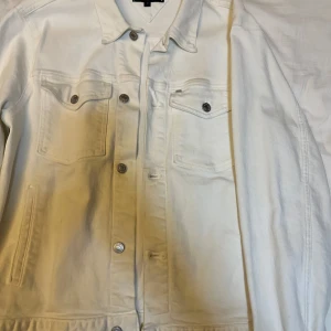 Vit jeansjacka från Tommy Hilfiger - Säljer en stilren vit jeansjacka från Tommy Hilfiger. Jackan har klassisk krage, knappar framtill och två bröstfickor med knapp. På baksidan finns en diskret logotyp i relief. Perfekt för en avslappnad look!