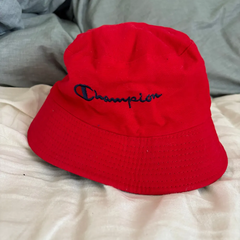 Snygg vändbar bucket hat från Champion i svart och rött. Ena sidan är svart med Champion-loggan i vitt och rött, medan den andra sidan är röd med Champion-text i blått. Perfekt för att variera din stil!. Asusteet.
