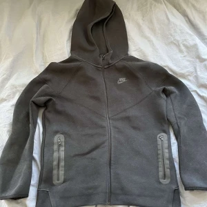 Grå hoodie från Nike - Säljer en svart/grå hoodie från Nike med dragkedja och huva. Tröjan har långa ärmar och två fickor med dragkedjor framtill. Perfekt för en avslappnad stil. Det är storlek xl i barn upplever den som storlek S. Skriv vid frågor eller funderingar😊