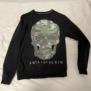 Svart tröja från Philipp Plein - Snygg svart tröja från Philipp Plein med ett stort dödskalletryck i kamouflagemönster på framsidan. Tröjan har långa ärmar och en rund halsringning. Perfekt för en cool och avslappnad stil. Pris sänks vid snabbaffär.