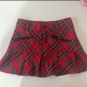 Rutig röd kortkjol med dragkedjor - Snygg rutig kortkjol i rött med svarta och vita detaljer. Kjolen har dragkedjor framtill och en liten ficka bak. Perfekt för en trendig look!