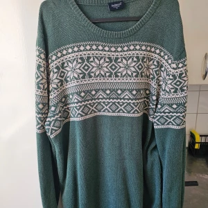 Grön stickad tröja från Redwood - Säljer en grön stickad tröja från Redwood med ett snyggt vitt mönster över bröstet och ärmarna. Perfekt för kyliga dagar med sin mjuka och bekväma passform. Tröjan har långa ärmar och rund halsringning.