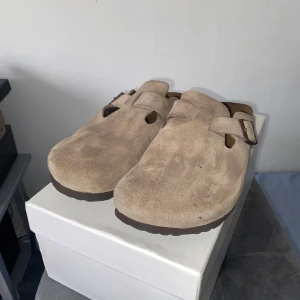 Birkenstock Boston - || Säljer ett par bekväma beige tofflor från Birkenstock i mocka. || De har en justerbar rem med spänne och en ergonomisk sula för extra komfort. || Perfekta för avslappnade dagar. || Använda under en sommar, så lite sletna med smuts här å där, men inget som man direkt lägger märke till. || Låda tillkommer EJ! || Storlek 45, passar som 44 ||
